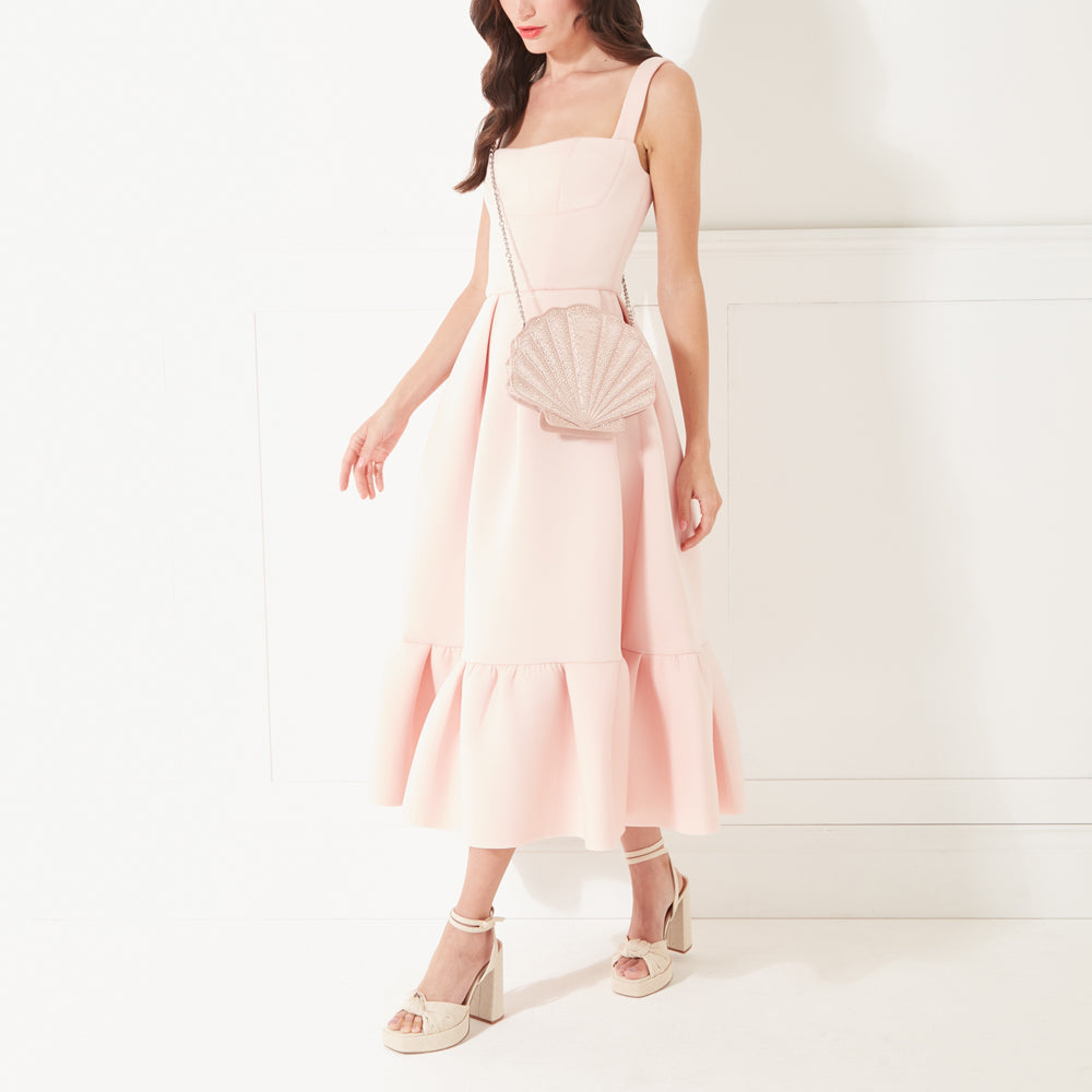 A full length image of a model with dark hair and red lipstick wearing a pale pink dress, heels and a blush pink satin shell shaped clutch bag on her shoulder.