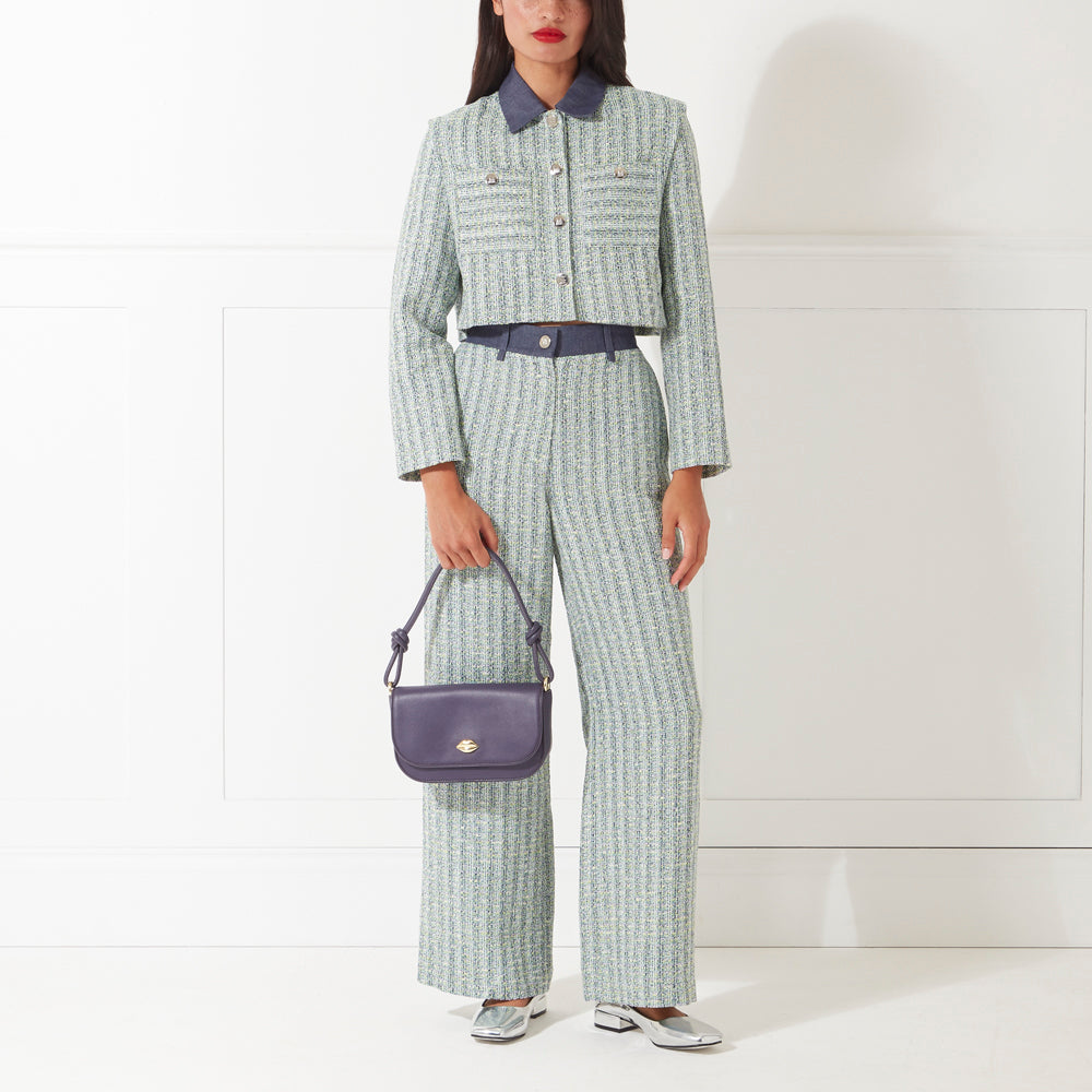A woman with red lipstick stands against a white background wearing a light green tweed pantsuit and silver shoes. She carries a dark purple handbag.