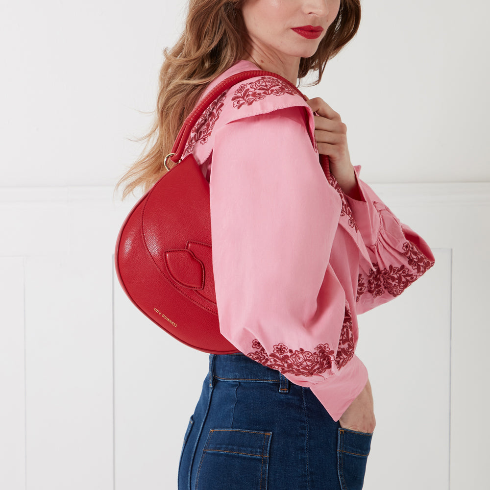 A red Lulu Guinness handbag with a lip design, held by a woman wearing a pink embroidered blouse and blue jeans against a white background.
