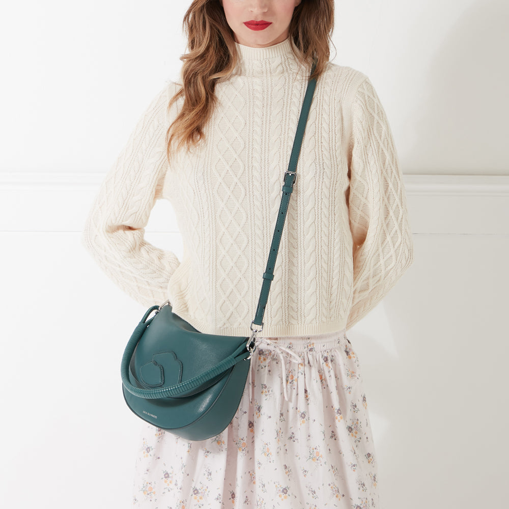 Woman wearing cream cable knit sweater and floral skirt, carrying a dark teal crossbody bag.
