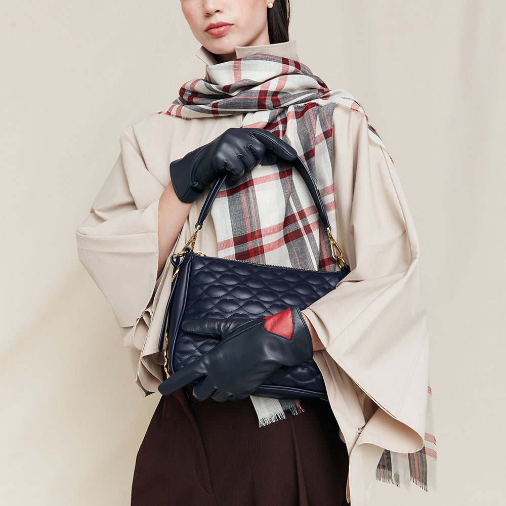 Person holding a navy quilted handbag wearing navy leather gloves with a red accent, wearing a beige coat and plaid scarf.