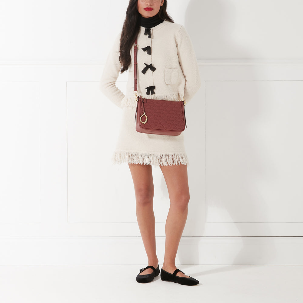 A woman in a cream-colored fringed skirt and cardigan with black bows, carries a russet crossbody bag and wears black Mary Jane shoes against a white background.
