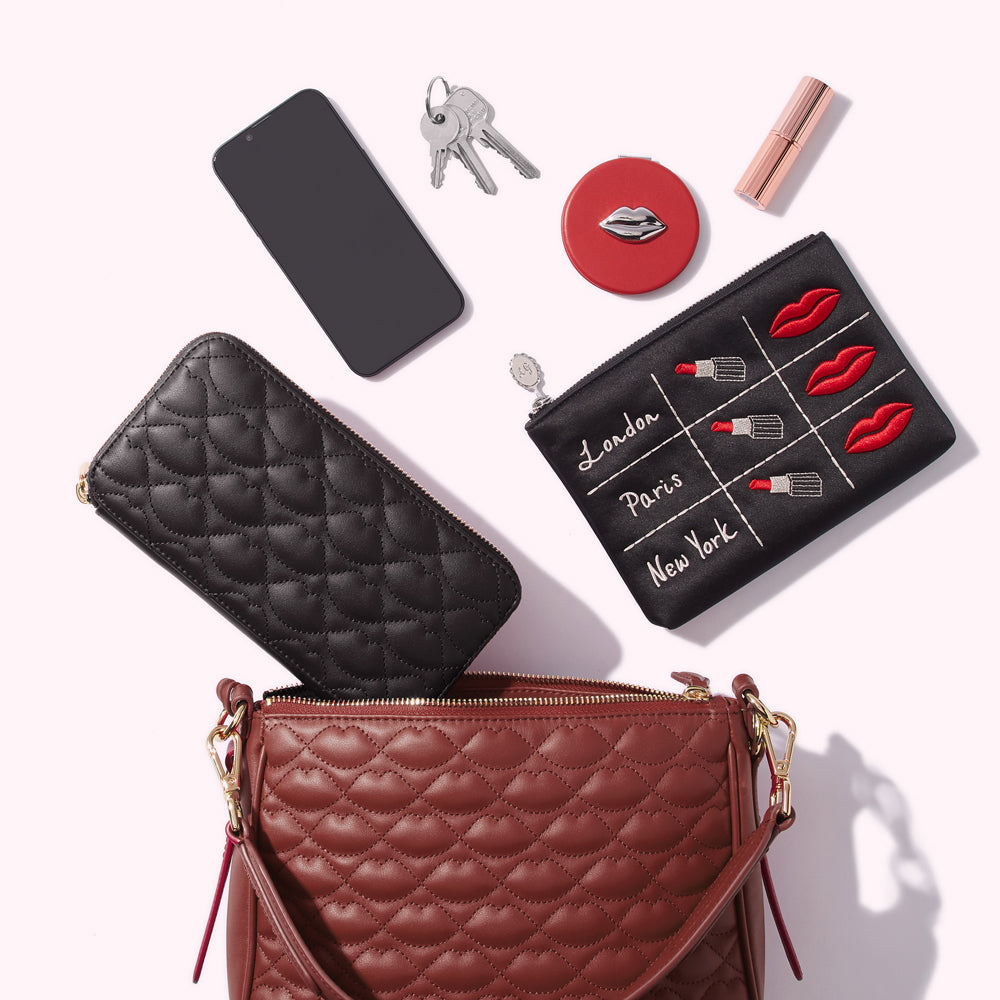 A russet quilted handbag with a black quilted wallet, smartphone, keys, compact mirror, lipstick, and a black quilted makeup bag inside, all on a white background.
