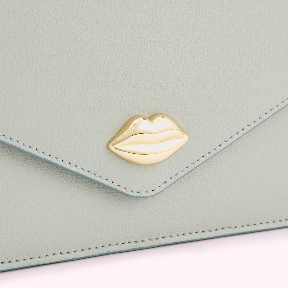 Close-up of a light sage green leather wallet with a gold lip-shaped clasp.
