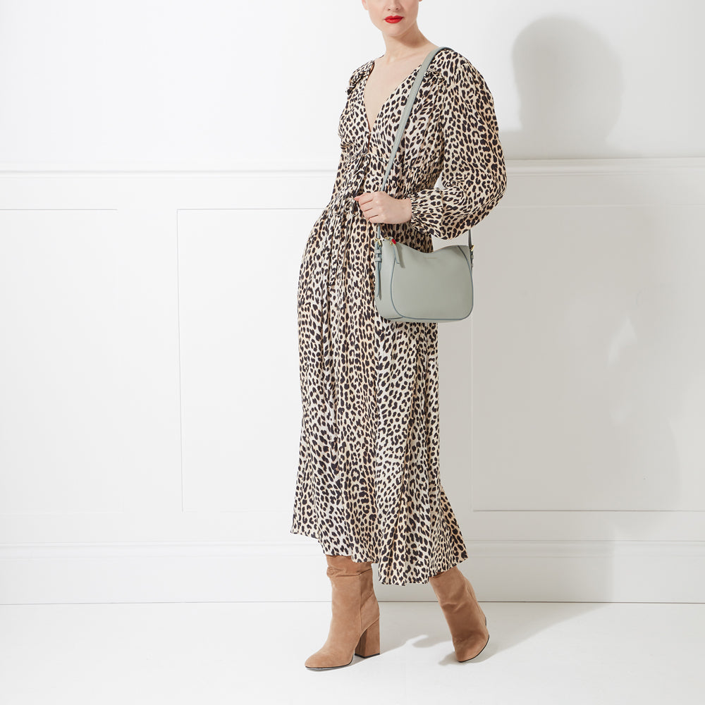A woman in a leopard print dress and tan boots carries a light sage green crossbody bag against a white background.

