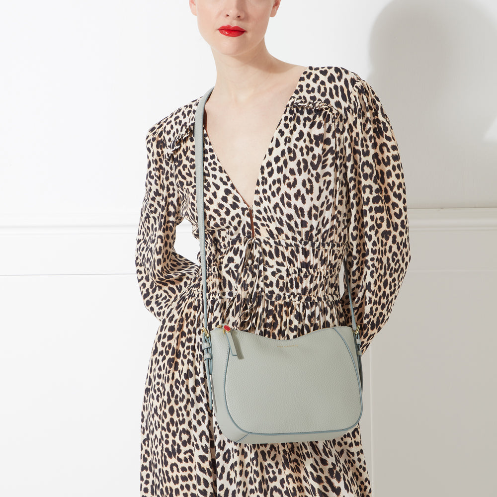 A light teal crossbody bag worn by a model in a leopard print dress against a white background.