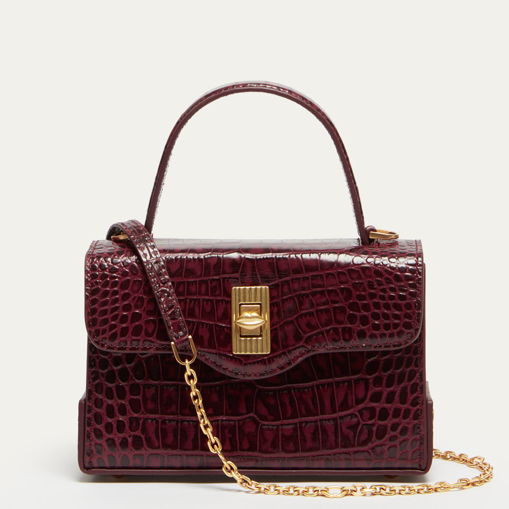 A structured burgundy croc-effect embossed leather handbag with a top handle, gold clasp, and a gold chain strap, photographed against a clean off-white background.
