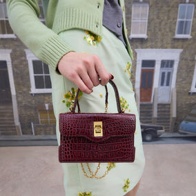 A women in a green cardigan and skirt holding a burgundy croc-effect leather handbag standing on a street.