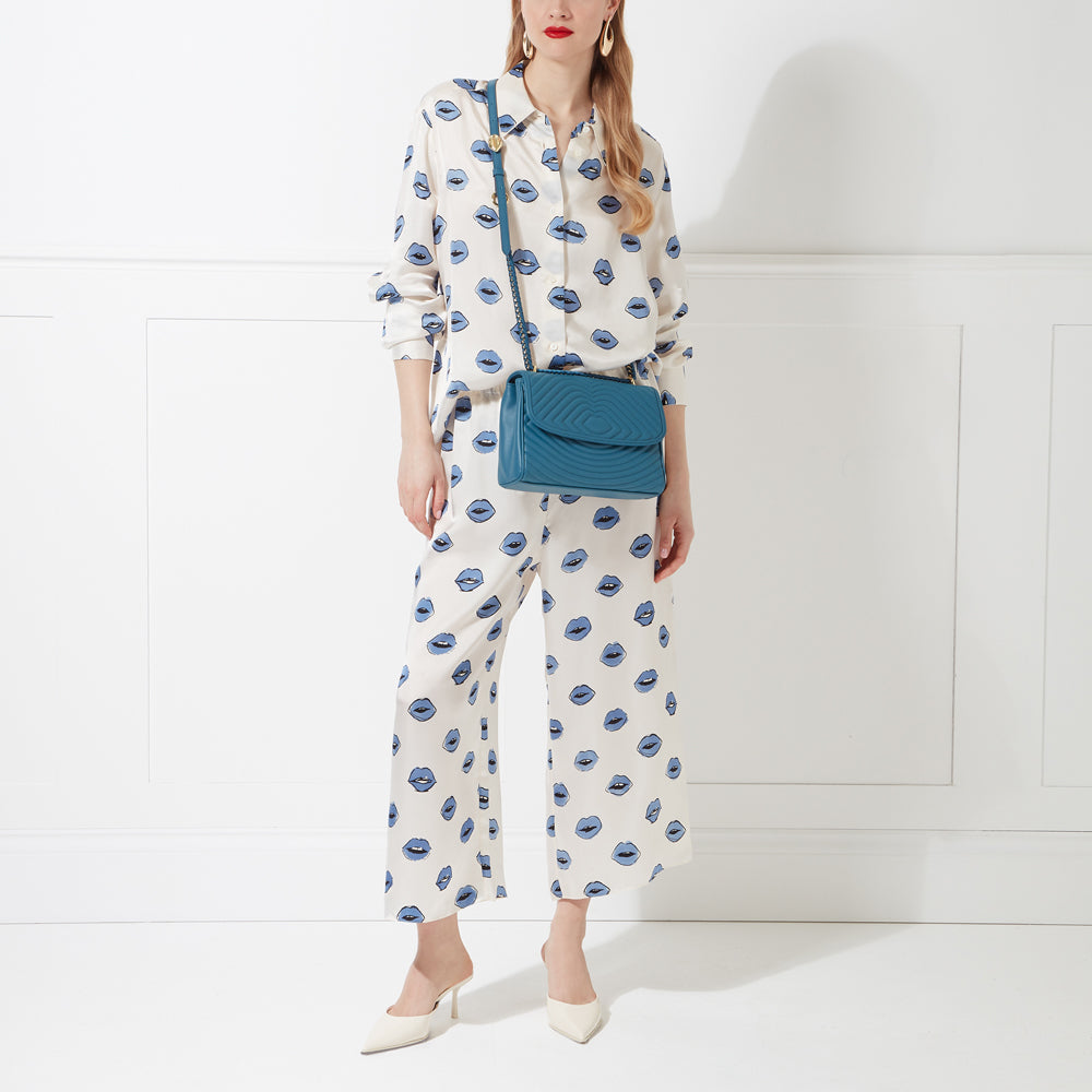 Woman in lip-print pants and shirt, wearing teal crossbody bag and white heels against a white background.

