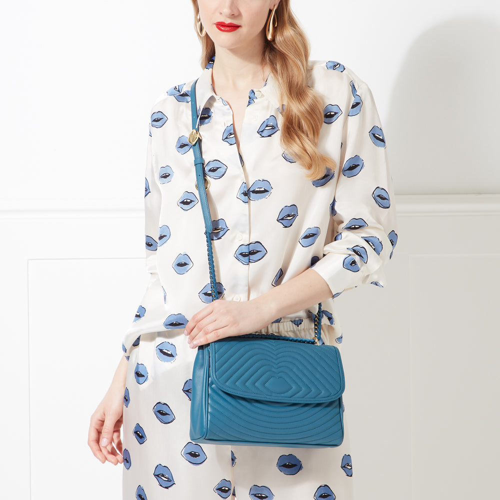 A woman with blonde hair is wearing a cream-colored shirt with a blue lips pattern and teal quilted handbag with a gold chain strap.