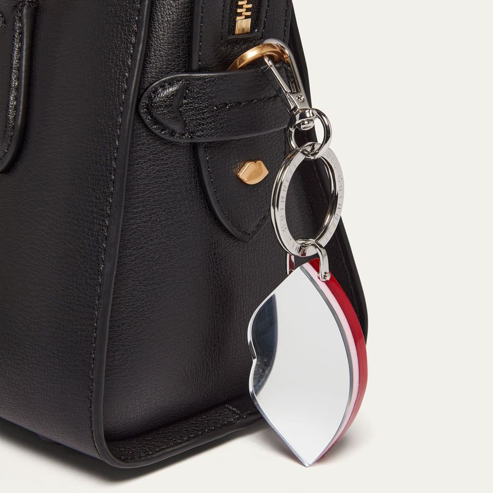 A silver mirror keychain featuring a flat, lip-shaped charm, attached to a lobster clasp and rings, shown clipped onto a black leather handbag.