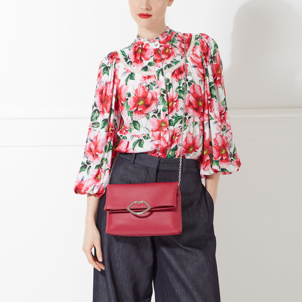 Woman wearing a floral blouse and dark pants, carrying a red crossbody bag with a silver lip-shaped clasp.
