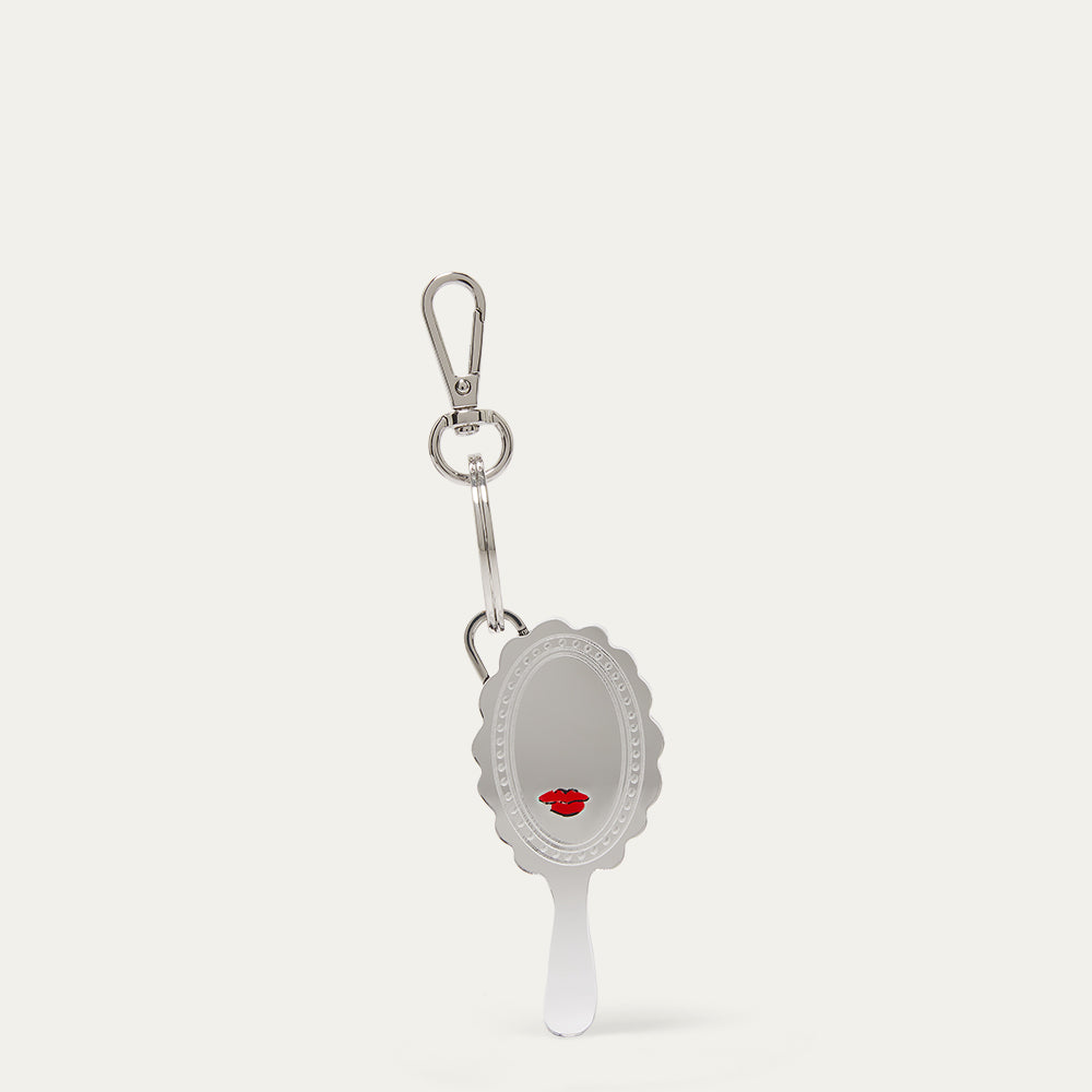 A silver hand mirror keychain with a small red lips graphic in the lower half, attached to a silver carabiner on a light beige background.