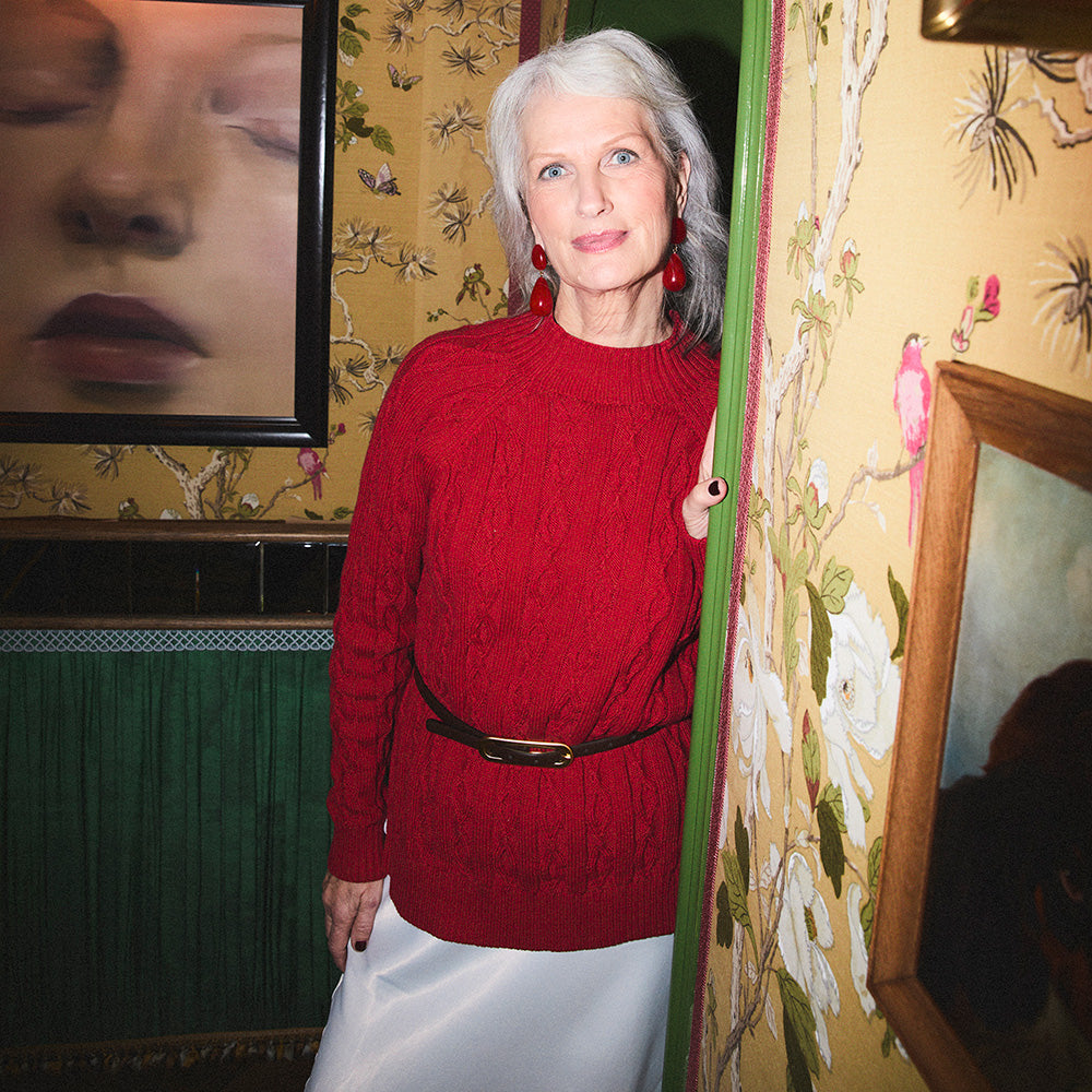 Woman in a red sweater standing in a room with floral wallpaper and framed pictures.