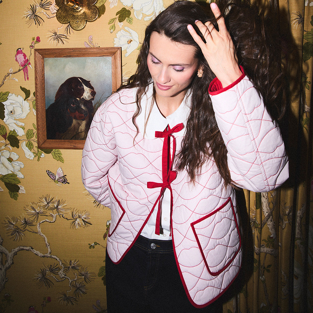 Woman wearing a pink quilted jacket with red lip shaped quilting and trim in a room with floral wallpaper.
