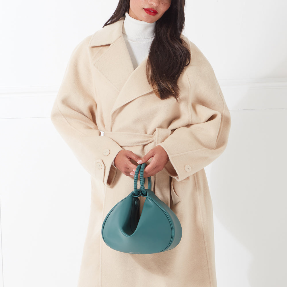 Woman in beige coat holding teal handbag.
