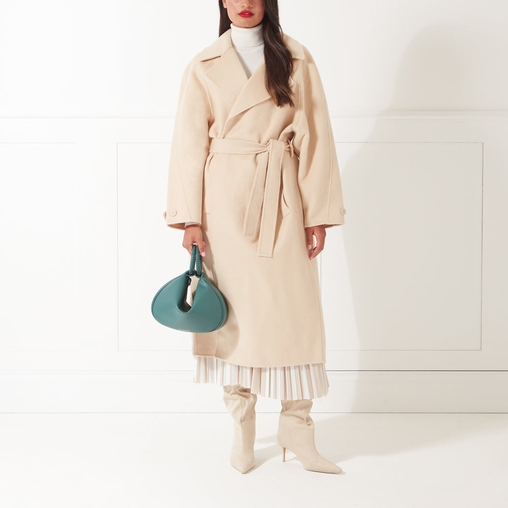 Woman in beige coat, teal handbag, and beige boots against a white background.
