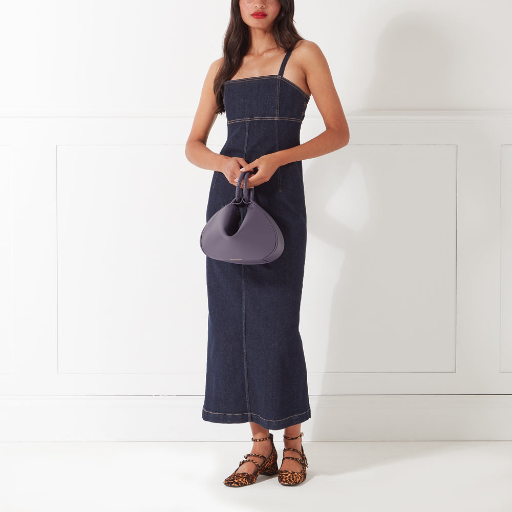 Woman in a denim dress holding a purple handbag and wearing leopard print shoes against a white background.
