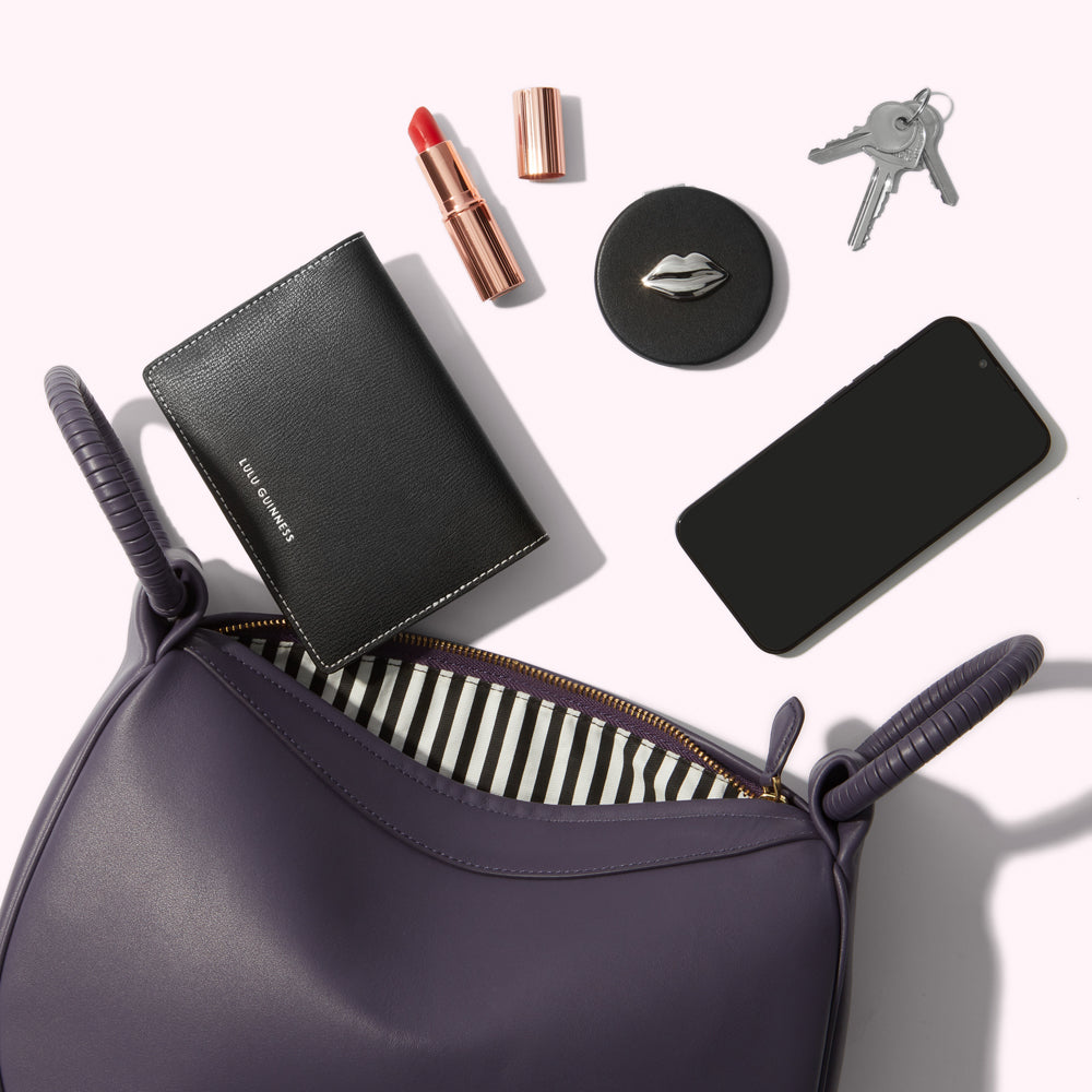 A purple handbag is open to reveal a black wallet, lipstick, compact mirror, and smartphone. Keys are also on the white background.
