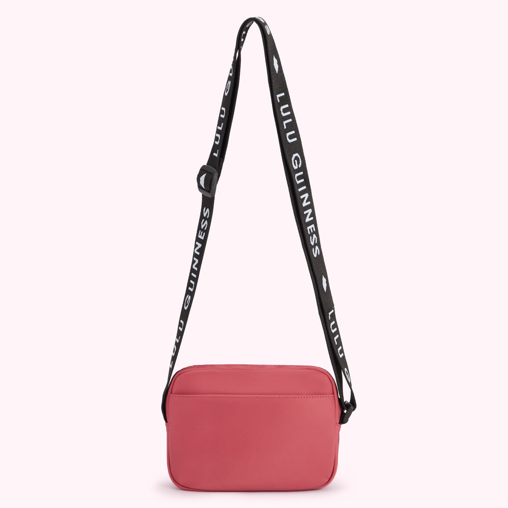 Back view of a coral Lulu Guinness crossbody bag with a black strap that has the brand name printed on it.
