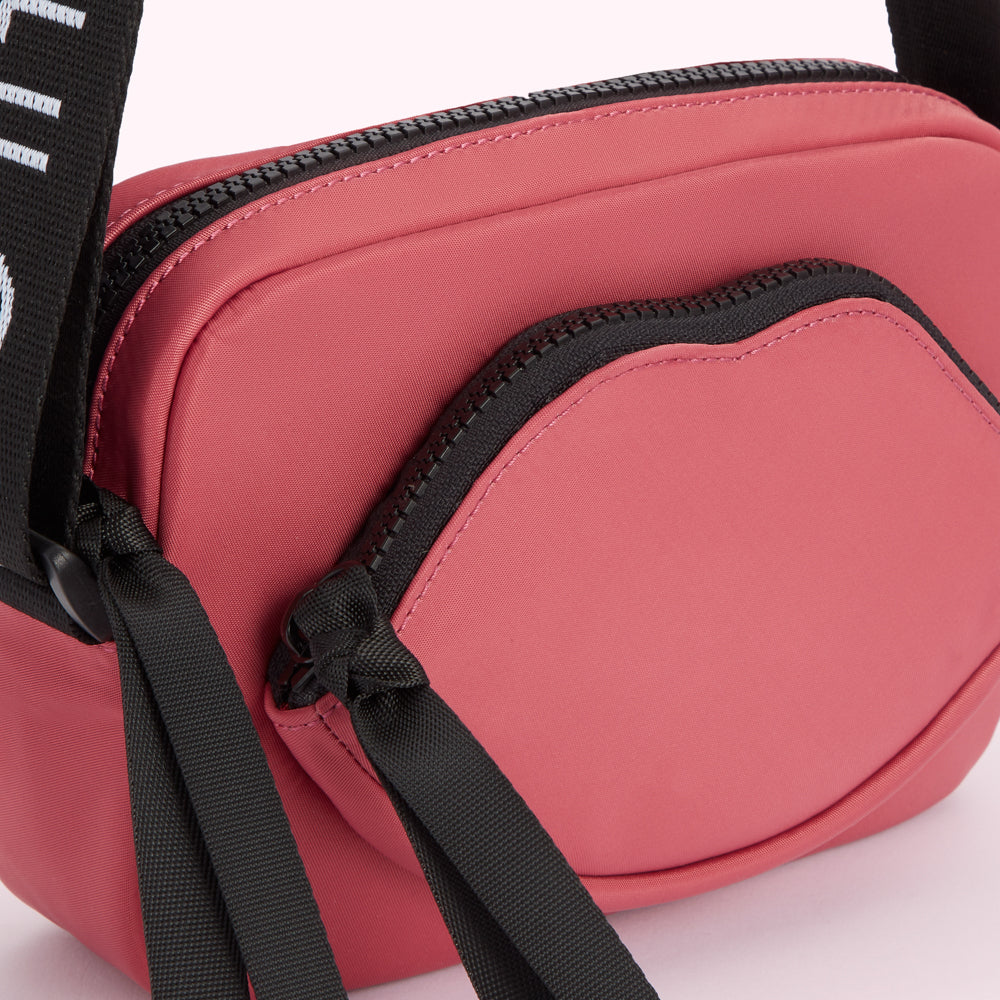 Dusty rose pink crossbody bag with a lip-shaped front pocket and black straps.
