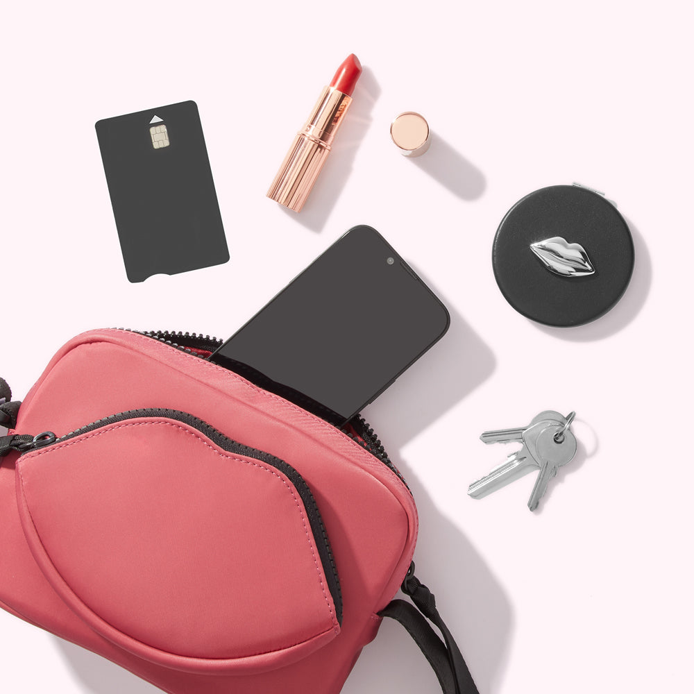 A coral-coloured crossbody bag with a lip-shaped pocket containing a smartphone, credit card, lipstick, compact mirror, and keys.