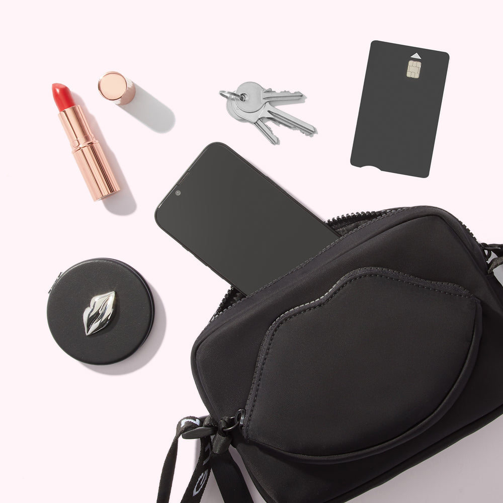 A black crossbody bag with a lip-shaped detail lies open on a white background, spilling its contents: a smartphone, keys, a credit card, lipstick, and a compact mirror.