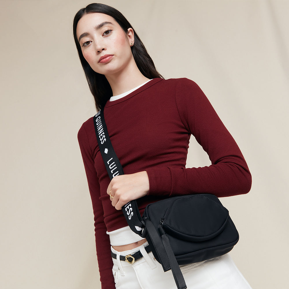 Woman holding a black crossbody bag with 'Lulu Guinness' branding on the strap against a beige background