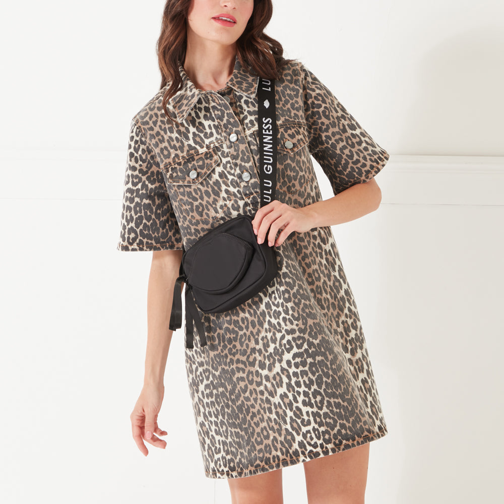 A model with dark hair wearing a leopard print denim dress and a black nylon crossbody bag with a lip shaped pocket and 'Lulu Guinness' branded strap.