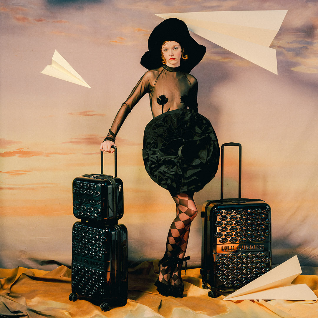 Woman in black outfit with patterned stockings standing between two black suitcases against a sunset sky.
