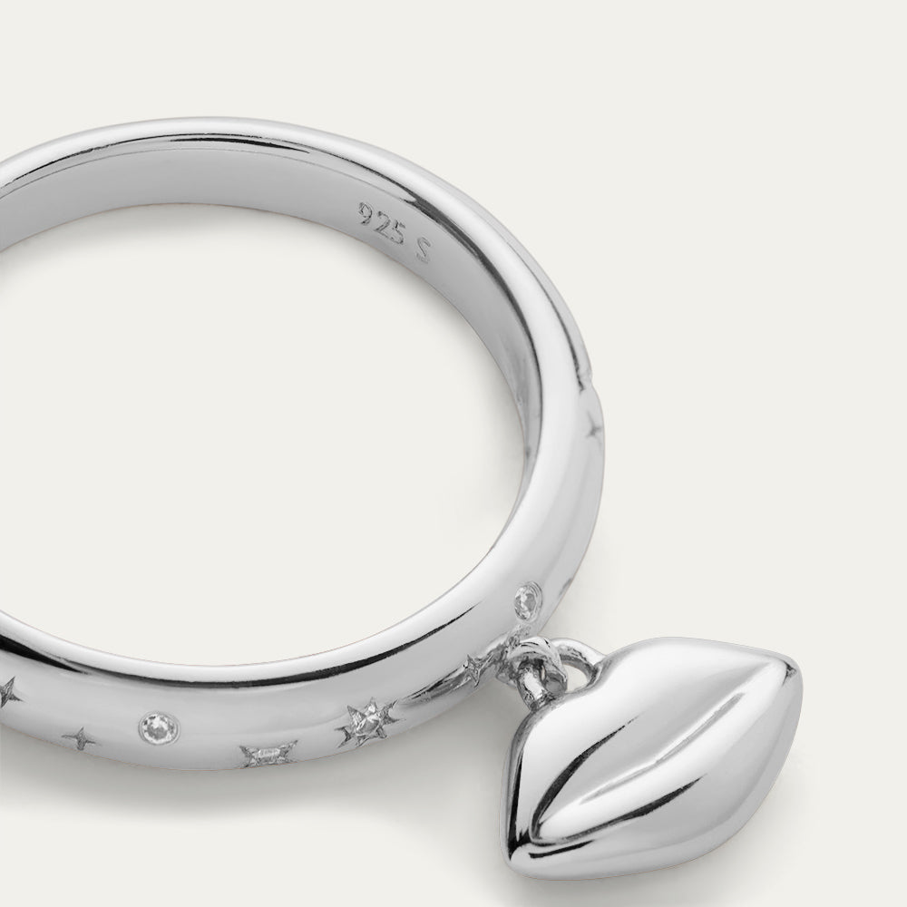 Close-up image of a silver ring with a lip-shaped pendant and inset stones on a off white background.
