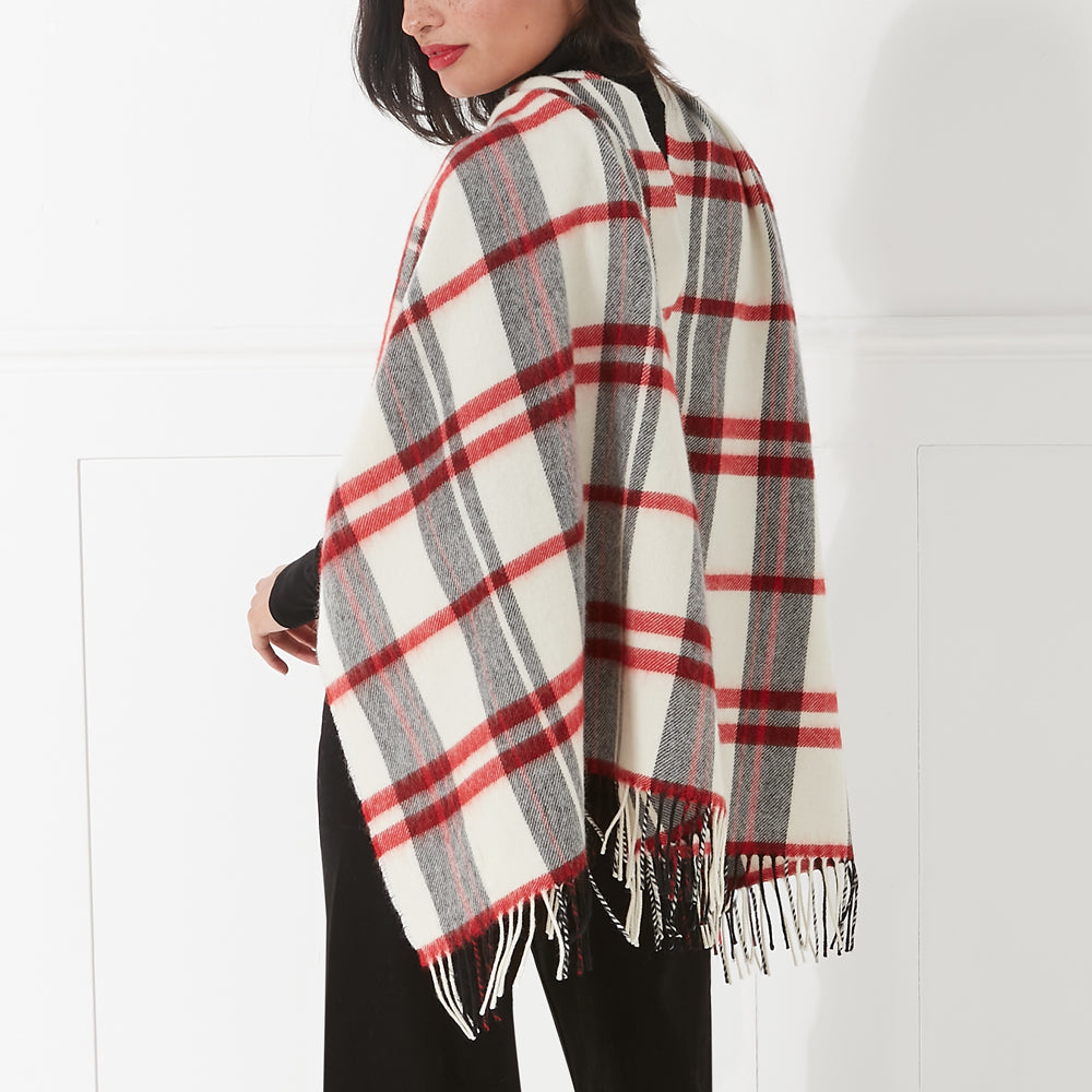 Woman wearing a cream, red, and grey plaid or tartan poncho with black pants against a white background.
