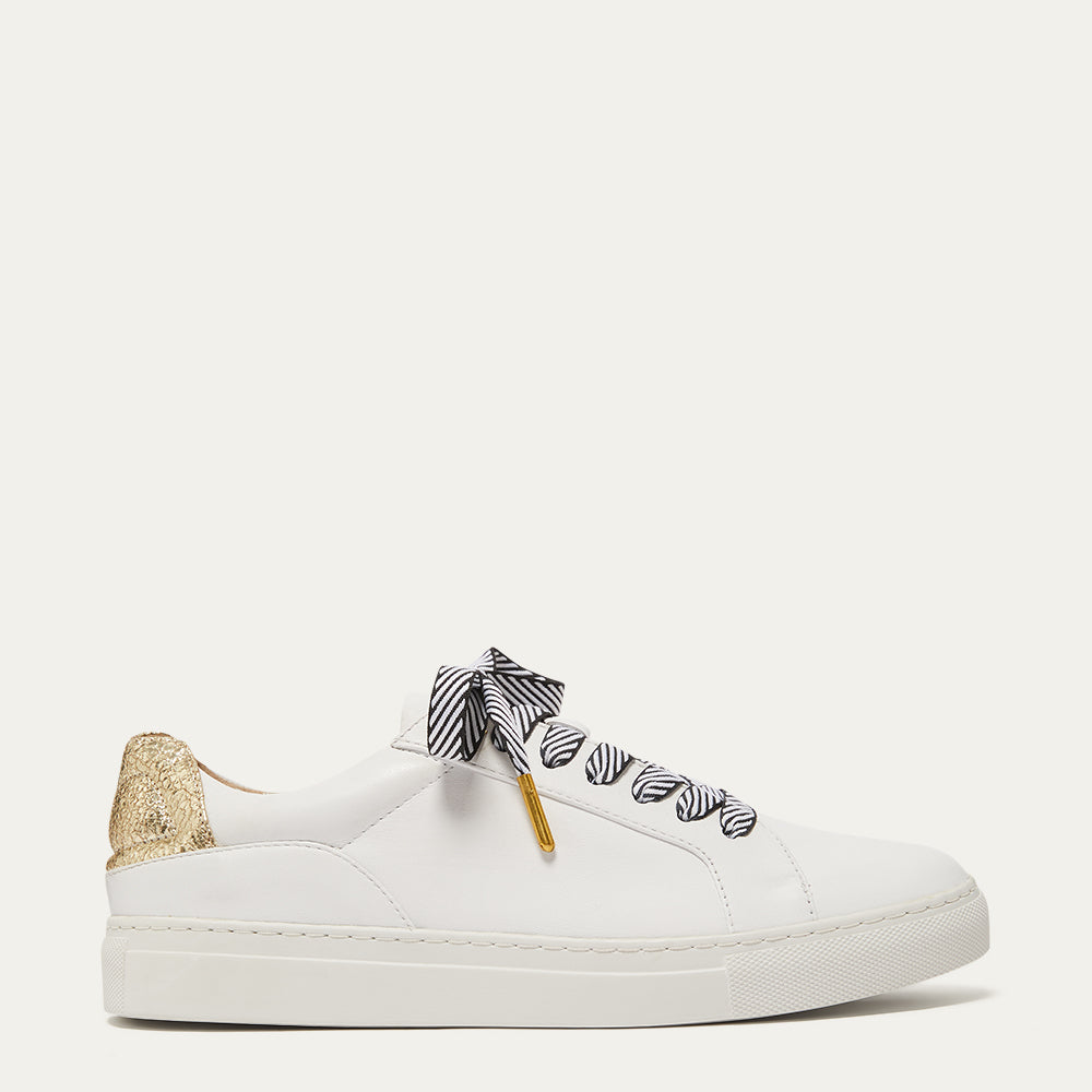 A white leather low-top sneaker with a gold heel tab, black and white patterned laces tied in a bow, and a small gold tag, shown against a solid off-white background.