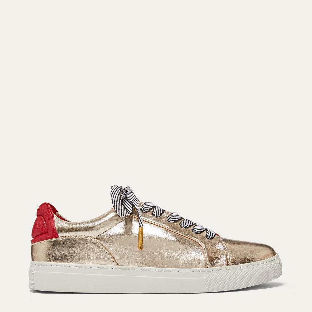 A metallic gold leather low-top sneaker with a red heel tab, striped black-and-white laces, and a white rubber sole, shown from a side profile on a light beige background.