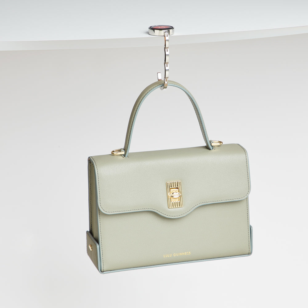 A sage green Lulu Guinness handbag hangs from a silver hook attached to a white table.