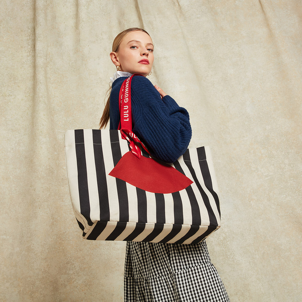 Woman holding a black and beige striped tote bag with a red lip printed on the front against a beige background