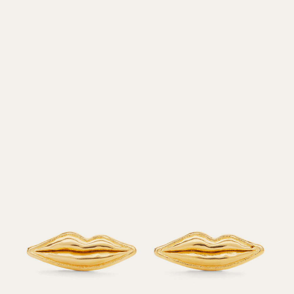 A pair of gold lips-shaped stud earrings on a light cream background.