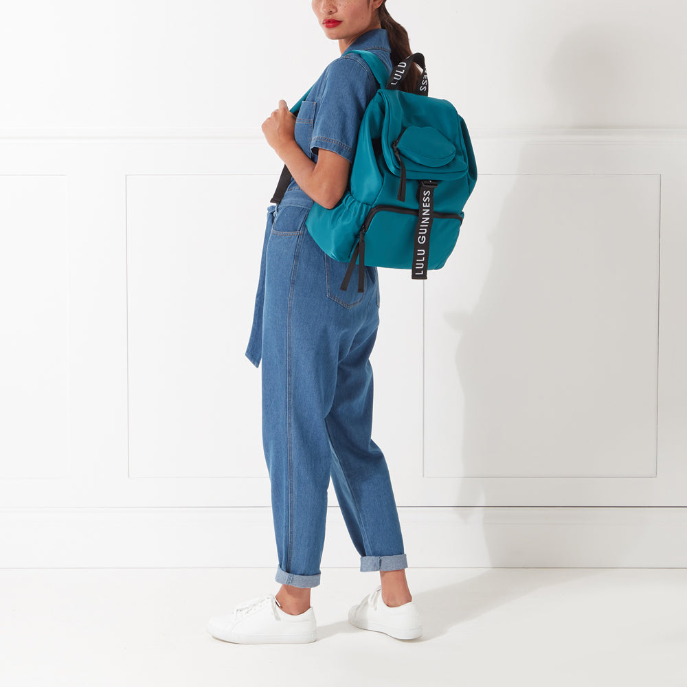 Woman wearing denim overalls and white sneakers with teal Lulu Guinness backpack.
