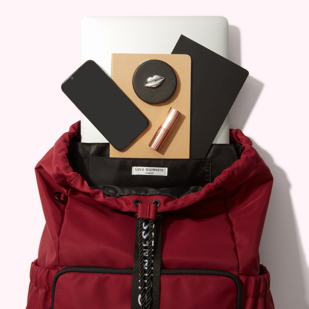 A burgundy Lulu Guinness backpack with a laptop, phone, notebooks, lipstick, and compact mirror inside.
