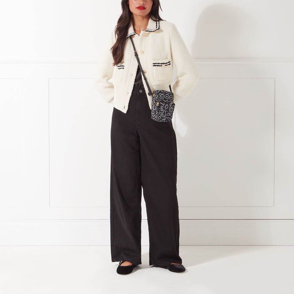 Woman wearing a white tweed jacket, black wide-leg trousers, black flats, and a black crossbody bag against a white background.
