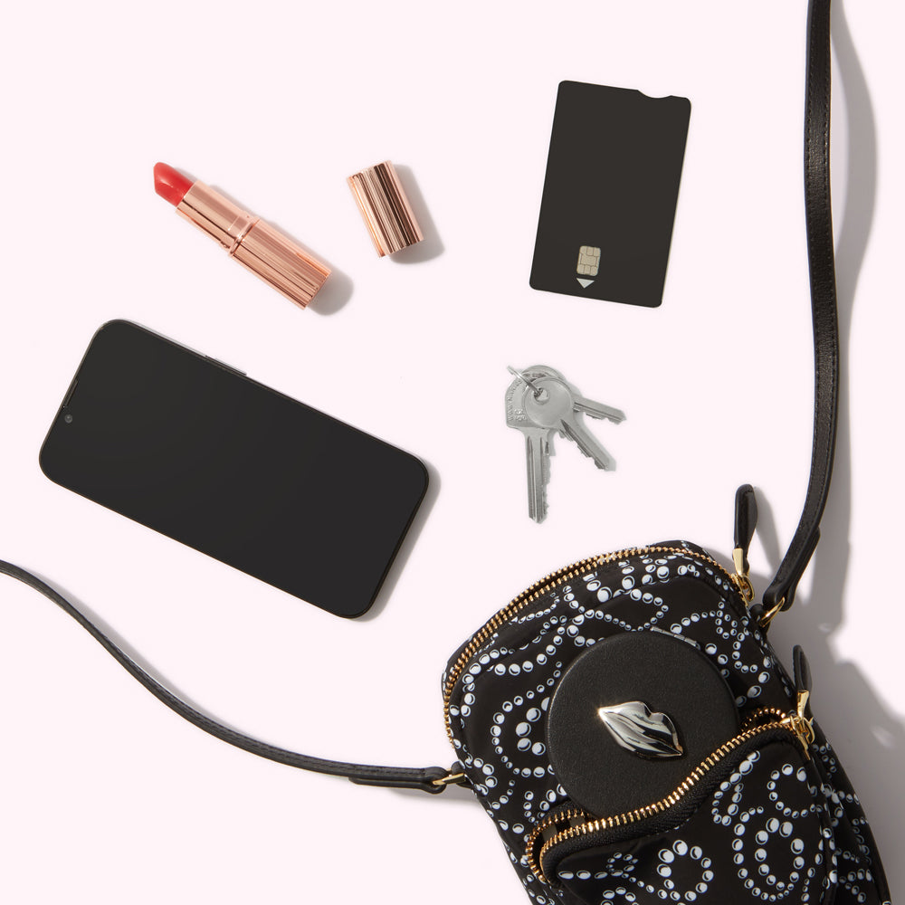 A black crossbody bag with a white pearl pattern lies on a white background next to a smartphone, keys, lipstick, and a credit card.