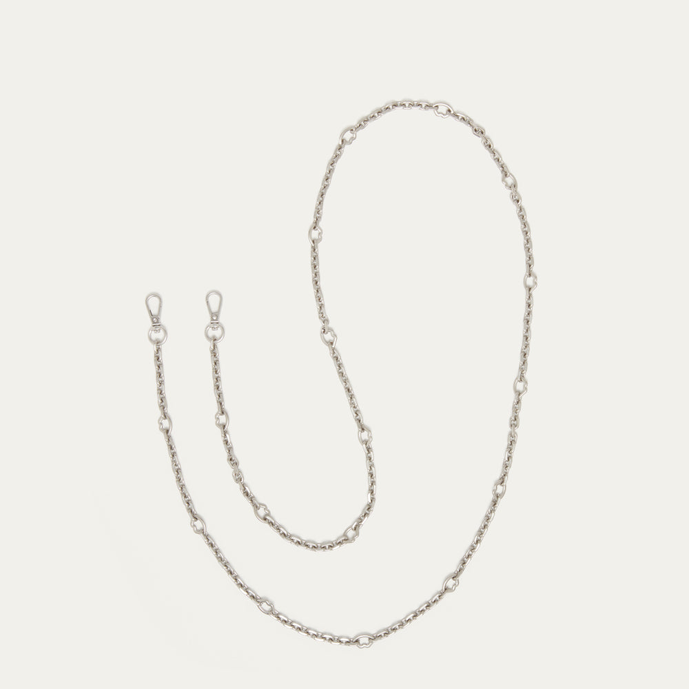 A silver chain accessory with alternating small oval links and larger lip-shaped links, laid out in a loose loop on a light cream background.