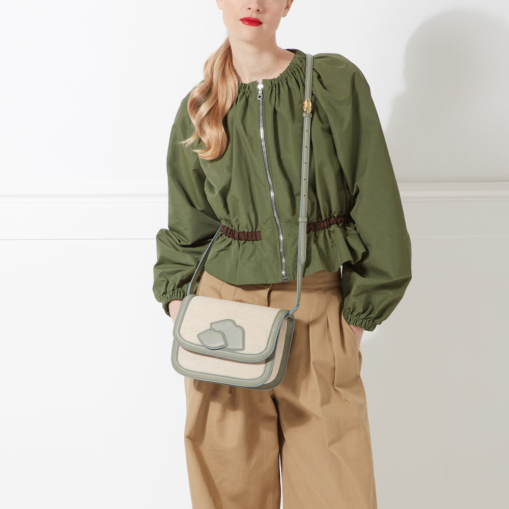 Woman wearing olive green zip-up jacket and khaki pants, carrying a light beige crossbody bag with sage green lip-shaped details against a white background.
