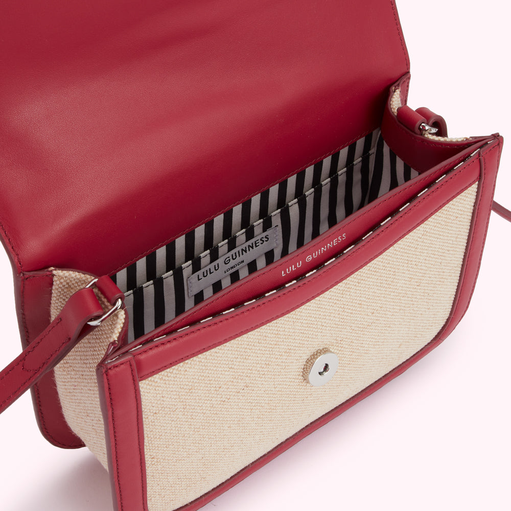 Open Lulu Guinness crossbody bag with red leather trim and beige canvas body. The interior lining is black and white striped.
