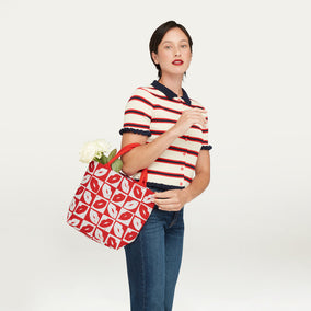 A woman wearing a red, white, and navy striped short-sleeve polo shirt and blue jeans holds a red and white checkerboard lip patterned tote bag with white flowers tucked inside, posed against a solid off-white studio background.