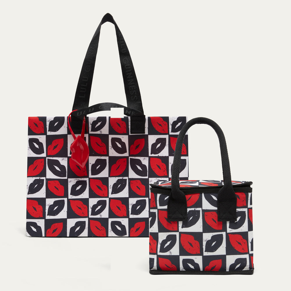 Two tote bags with black handles and a repeating lip print on a white, black, and red checkered background, shown on a light gray studio background.