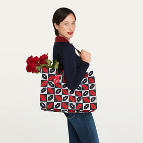 A woman wearing a navy long-sleeve top and blue jeans carries a large tote bag with a black and white checkerboard pattern of red and black lips. She is holding red roses in one of the bag's handles against a plain off-white background.