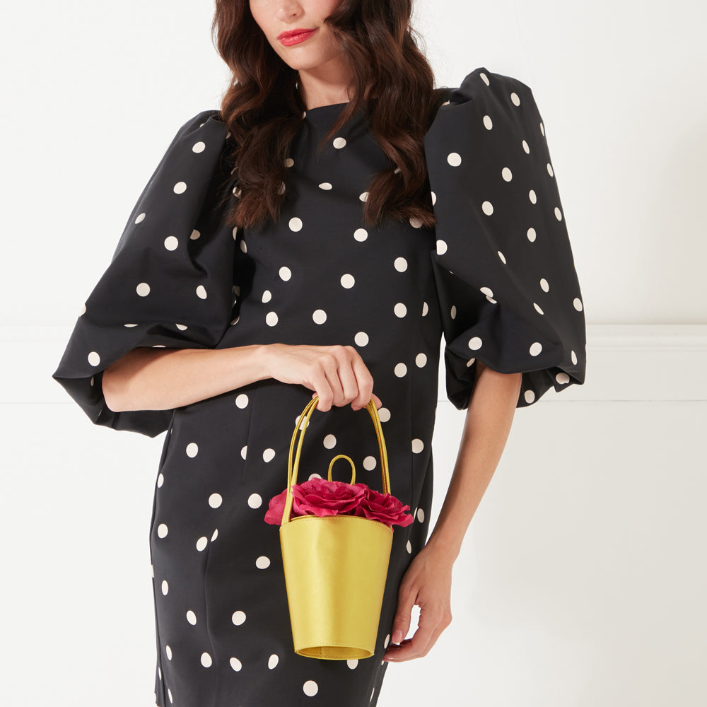 A full length image of a model wearing a polka dot dress and black heeled shoes, holding a chartreuse and pink rose basket bag.