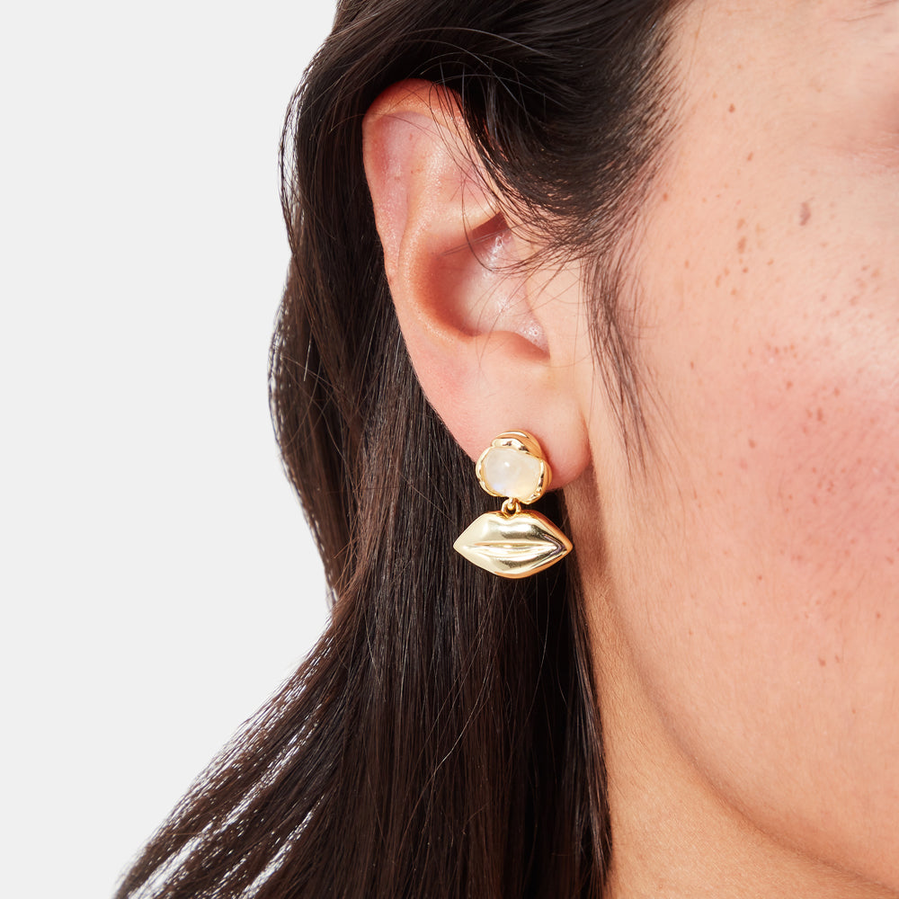 Close-up of a woman's ear wearing gold lip-shaped earrings with a moonstone accent.
