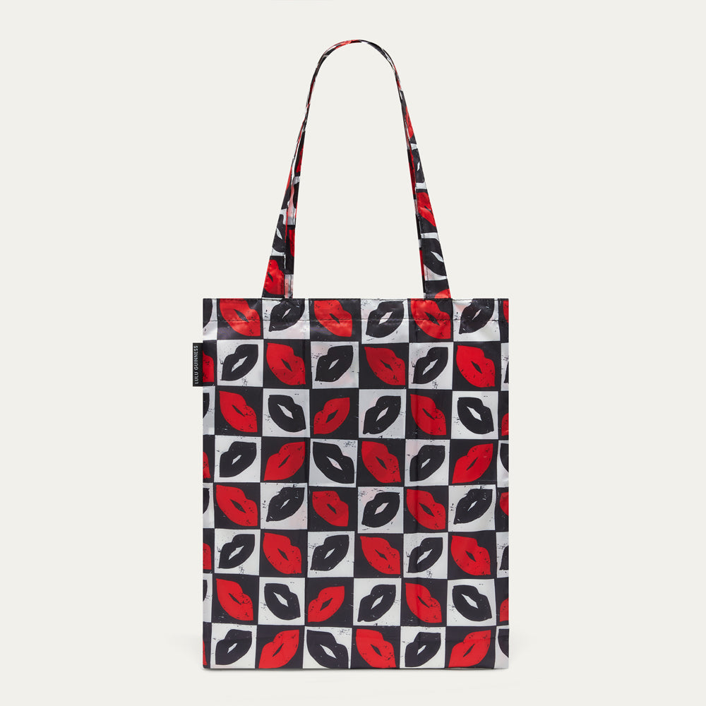 A tote bag with a repeating pattern of red and black lips on a white and black checkerboard background, featuring dual shoulder straps, shown against a light gray background.
